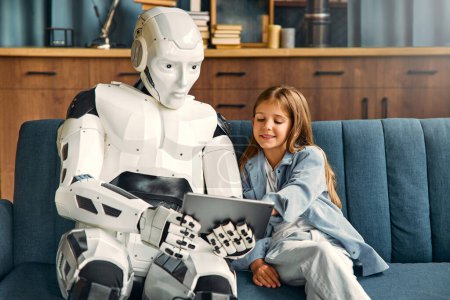 Téléchargez les photos : Robot femme de ménage infirmière en utilisant une tablette avec enfant fille assise sur le canapé dans le salon. Collaboration entre les humains et l'intelligence artificielle. - en image libre de droit