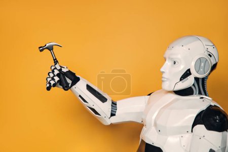 Téléchargez les photos : Robot tenant un marteau dans sa main isolé sur un fond orange. Concept d'un maître robot. Collaboration entre les humains et l'intelligence artificielle. - en image libre de droit