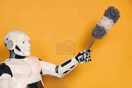 Téléchargez les photos : ROBOT tenant un pipidastra nettoyant la poussière dans sa main isolé sur un fond orange. Concept d'un robot femme de ménage. Collaboration entre les humains et l'intelligence artificielle. - en image libre de droit