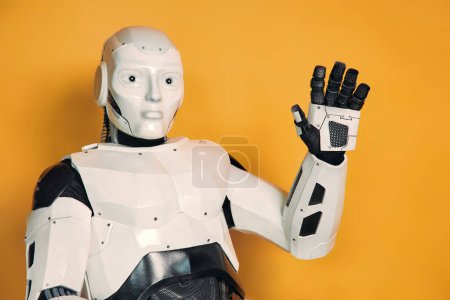Téléchargez les photos : Robot levant la main en saluant isolé sur fond orange. Collaboration entre les humains et l'intelligence artificielle. - en image libre de droit