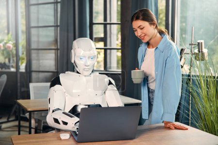 Foto de Mujer joven con una taza de café y un robot humanoide trabajando mientras está sentado en un ordenador portátil en una oficina moderna. Colaboración entre humanos e inteligencia artificial. - Imagen libre de derechos