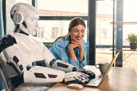 Téléchargez les photos : Jeune femme avec une tasse de café et un robot humanoïde travaillant assis sur un ordinateur portable dans un bureau moderne. Collaboration entre les humains et l'intelligence artificielle. - en image libre de droit