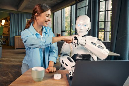 Téléchargez les photos : Jeune femme avec une tasse de café et un robot humanoïde travaillant assis à un ordinateur portable dans un bureau moderne, poing à poing geste. Collaboration entre les humains et l'intelligence artificielle. - en image libre de droit