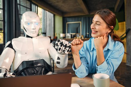 Young woman with a cup of coffee and a humanoid robot working while sitting at a laptop in a modern office, fist to fist gesture. Collaboration between humans and artificial intelligence.