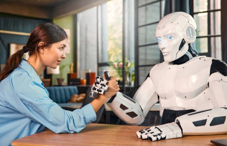 Photo for Young woman and humanoid robot doing arm wrestling while sitting at a table in a modern office. Collaboration between humans and artificial intelligence. - Royalty Free Image