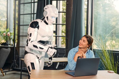 Foto de Mujer joven y un robot humanoide trabajando mientras está sentado en una computadora portátil en una oficina moderna. Colaboración entre humanos e inteligencia artificial. - Imagen libre de derechos