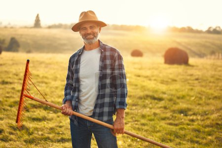 Téléchargez les photos : Un beau fermier mûr debout avec une fourche sur le fond d'un champ lors d'une chaude journée de travail dans une ferme. Concept d'agriculture et d'agriculture. - en image libre de droit