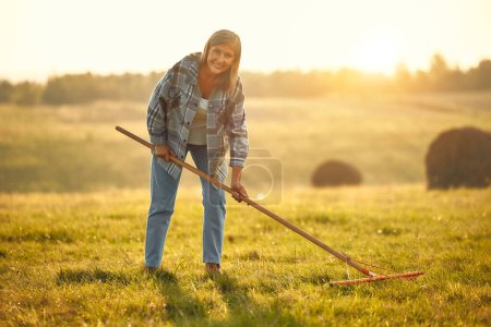 Téléchargez les photos : Mature belle agricultrice debout avec une fourche sur le fond d'un champ par une chaude journée de travail sur une ferme. Concept d'agriculture et d'agriculture. - en image libre de droit