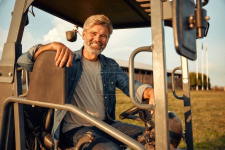 Téléchargez les photos : Un beau fermier mature assis dans un tracteur sur un champ par une chaude journée de travail dans une ferme. Concept d'agriculture et d'agriculture. - en image libre de droit