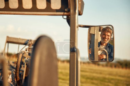 Foto de Granjero macho guapo maduro sentado en un tractor en un campo en un día caluroso trabajando en una granja. Concepto de agricultura y agricultura. - Imagen libre de derechos