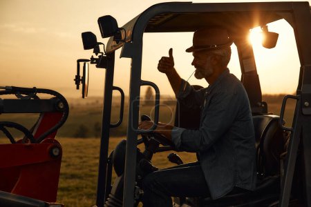 Foto de Maduro granjero macho guapo en un sombrero sentado en un tractor en un campo al atardecer después de un duro día de trabajo. Concepto agricultura y agricultura. - Imagen libre de derechos