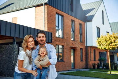 Photo for Moving of a young family to a new house. Couple with child standing in front of their newly bought house. Renting and buying a home. - Royalty Free Image