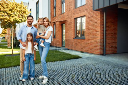 Photo for Moving of a young family to a new house. Couple with two kids daughters standing in front of their newly bought house. Renting and buying a home. - Royalty Free Image