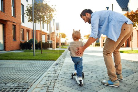 Téléchargez les photos : Papa apprend à sa petite fille mignonne à monter un scooter pour enfants dans la cour près de la maison. Papa jouant avec sa fille dans la rue. - en image libre de droit