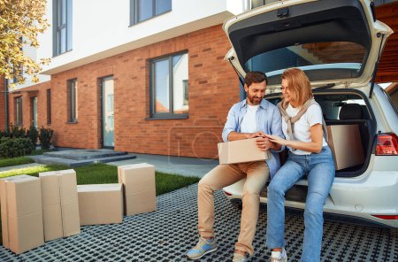 Photo for Young married couple sitting in the trunk of a car and unloading luggage in boxes to their new home. Renting and buying a home. Moving concept. - Royalty Free Image