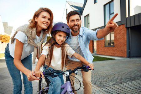 Photo for A happy father and mother taught his little daughter to ride a bicycle. A child learns to ride a bicycle. Family activities in summer. - Royalty Free Image