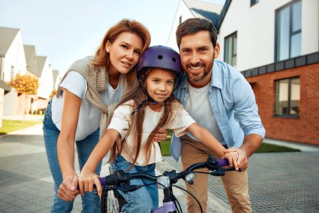Téléchargez les photos : Un père et une mère heureux ont appris à sa petite fille à faire du vélo. Un enfant apprend à faire du vélo. Activités familiales en été. - en image libre de droit