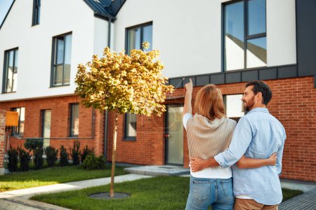 Foto de Una joven pareja casada feliz de pie cerca de una nueva casa y abrazándose. Alquilar y comprar una casa. Concepto móvil. - Imagen libre de derechos
