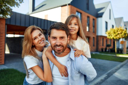 Photo for A young happy family with a child hugging each other are standing on the street near their new home. Renting and buying real estate. Moving concept. - Royalty Free Image