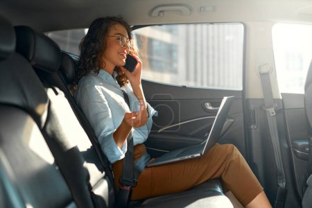 Photo for Attractive business woman working on her laptop at the back sit of a car and talking on the phone on the way to meeting. - Royalty Free Image