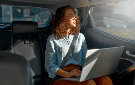 Foto de Young business woman with a modern laptop sitting in the back seat of luxury car. Successful startup, distance working, female boss concept. - Imagen libre de derechos