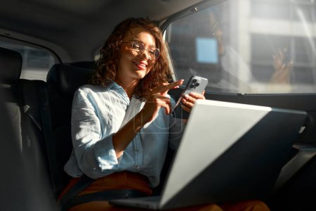 Téléchargez les photos : Belle femme d'affaires travaillant sur son ordinateur portable à l'arrière assis d'une voiture et parlant au téléphone sur le chemin de la réunion
. - en image libre de droit