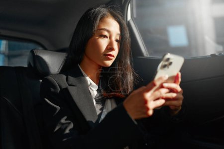 Foto de Exitosa joven empresaria asiática en ropa de oficina de moda trabajando con un teléfono inteligente en el asiento trasero del coche en la ciudad moderna urbana. - Imagen libre de derechos