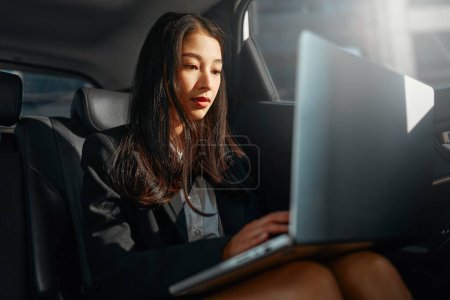 Photo for Young asian business woman with a modern laptop sitting in the back seat of luxury car. Successful startup, distance working, female boss concept. - Royalty Free Image