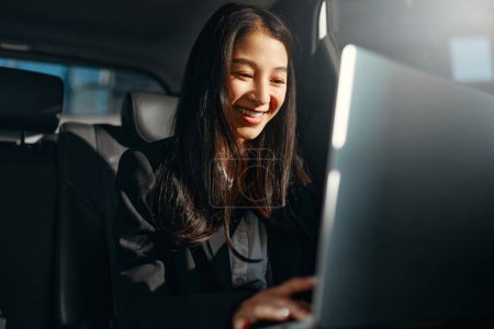 Photo for Young asian business woman with a modern laptop sitting in the back seat of luxury car. Successful startup, distance working, female boss concept. - Royalty Free Image