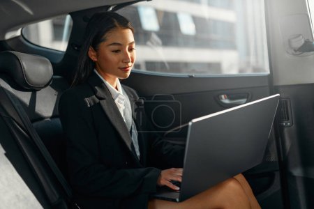 Téléchargez les photos : Jeune femme d'affaires asiatique avec un ordinateur portable moderne assis sur le siège arrière de la voiture de luxe. Démarrage réussi, télétravail, concept de boss féminin. - en image libre de droit