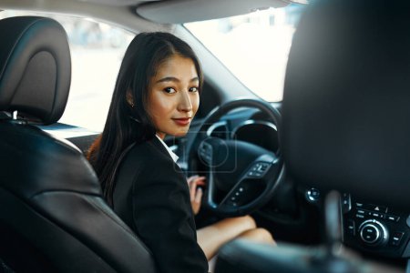 Téléchargez les photos : Belle asiatique jeune femme d'affaires souriante heureuse en costume noir au volant de sa nouvelle voiture. Acheter et louer une voiture. Voyages, tourisme, loisirs. - en image libre de droit