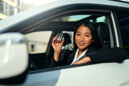 Téléchargez les photos : Portrait de belle femme d'affaires asiatique assise dans sa nouvelle voiture et montrant les clés. Acheter et louer une voiture. Voyages, tourisme, loisirs. - en image libre de droit