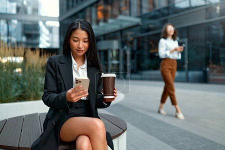 Foto de Una joven hermosa mujer de negocios asiática bebe café y usa un teléfono inteligente mientras está sentada en la calle contra el telón de fondo de los centros de negocios. - Imagen libre de derechos