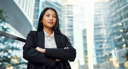 Photo for Beautiful Asian business woman in business clothes standing with her arms crossed on the steps against the backdrop of business centers. - Royalty Free Image
