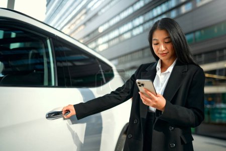 Photo for Beautiful young happy asian business woman in black suit standing near her new car and using smartphone. Buying and renting a car. Travel, tourism, recreation. - Royalty Free Image