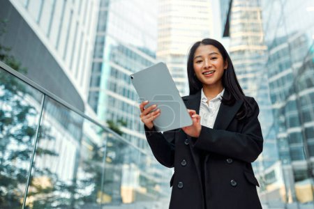 Photo for Asian business woman in a black suit holding a tablet while standing on the steps against the backdrop of business centers. - Royalty Free Image