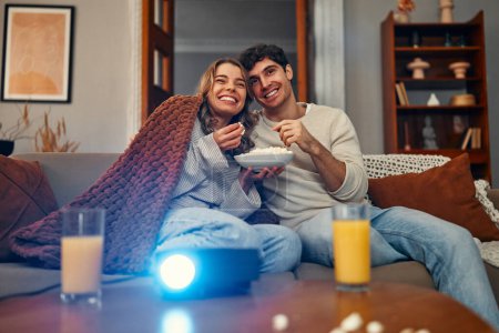 Téléchargez les photos : Joyeuse Saint-Valentin. Un jeune couple amoureux assis sur le canapé dans le salon à la maison, mangeant du pop-corn, couvert d'une couverture et regardant un film sur le projecteur. - en image libre de droit