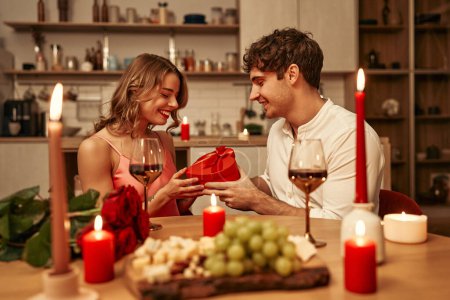 Photo for Happy Valentine's Day. A couple with glasses of wine by candlelight sitting in the kitchen at the table, romantically spending the evening together. A man giving his beloved a heart-shaped gift box. - Royalty Free Image