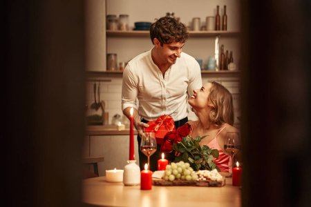 Téléchargez les photos : Joyeuse Saint-Valentin. Un couple avec des verres de vin aux chandelles assis dans la cuisine à la table, passer la soirée ensemble romantiquement. Un homme donnant à sa bien-aimée une boîte-cadeau en forme de coeur. - en image libre de droit