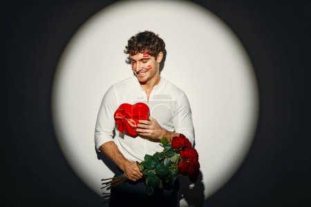 Téléchargez les photos : Joyeuse Saint-Valentin. Jeune homme sexy avec un bouquet de roses et un cadeau avec des traces de rouge à lèvres sur son visage de baisers, debout sur un fond blanc sous la lumière d'un projecteur. - en image libre de droit