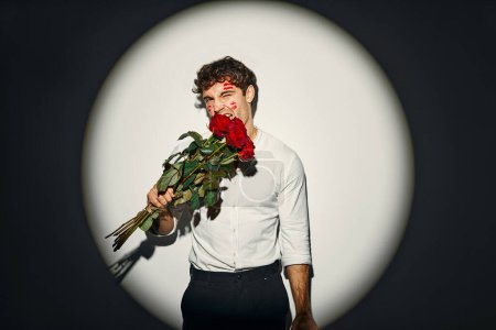 Téléchargez les photos : Joyeuse Saint-Valentin. Homme sexy avec un bouquet de roses avec des traces de rouge à lèvres en forme de lèvres sur son visage de baisers, debout sur un fond blanc sous la lumière d'un projecteur. - en image libre de droit