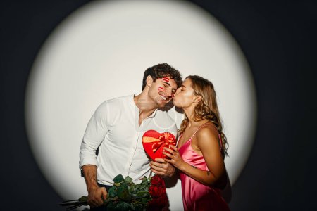 Téléchargez les photos : Joyeuse Saint-Valentin. Couple amoureux debout avec une boîte cadeau en forme de coeur et un bouquet de roses sur un fond blanc sous la lumière d'un projecteur. - en image libre de droit
