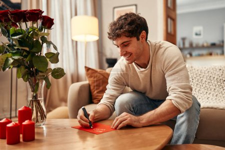 Foto de Feliz día de San Valentín. Un hombre sentado en una mesa en la sala de estar firma una tarjeta para su amada, felicitando a todos en el Día de San Valentín. Hay velas y un ramo de rosas sobre la mesa.. - Imagen libre de derechos