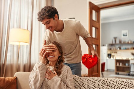 Photo for Happy Valentine's Day. A man gives a heart-shaped gift box to his beloved woman with his hand covering her eyes in the living room at home. Romantic evening together. - Royalty Free Image