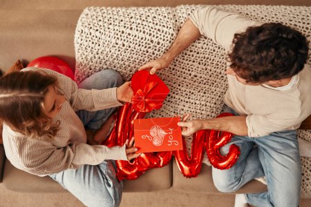 Photo for Happy Valentine's Day. Young couple in love exchanging gifts while sitting on the sofa in the living room at home. Woman giving a greeting card, man giving a heart-shaped gift box. - Royalty Free Image