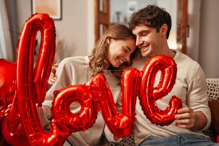 Foto de Feliz día de San Valentín. Pareja joven enamorada sosteniendo un globo con el amor de inscripción mientras está sentada en el sofá en la sala de estar en casa. Noche romántica juntos. - Imagen libre de derechos