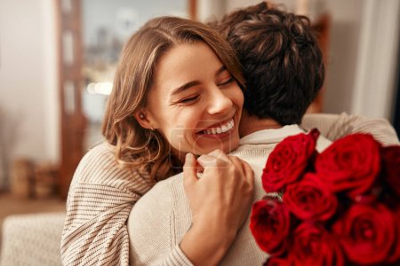Foto de Feliz día de San Valentín. Un hombre le da un ramo de rosas rojas a su amada mujer en la sala de estar de su casa, la mujer lo abraza tiernamente. Noche romántica juntos. - Imagen libre de derechos