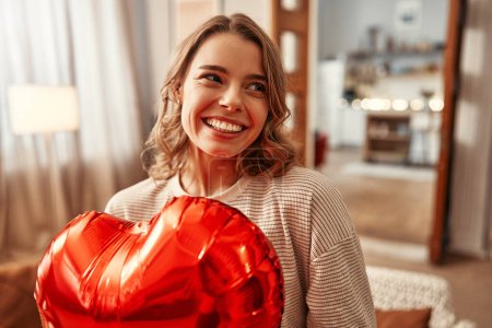 Téléchargez les photos : Joyeuse Saint-Valentin. Heureuse femme souriante avec ballon en forme de coeur rouge dans le salon à la maison. - en image libre de droit