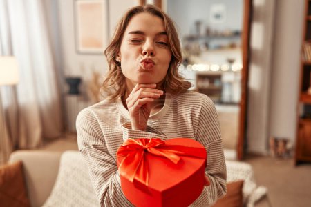 Téléchargez les photos : Joyeuse Saint-Valentin. Heureuse femme souriante tenant boîte cadeau en forme de coeur rouge avec arc dans le salon à la maison. - en image libre de droit