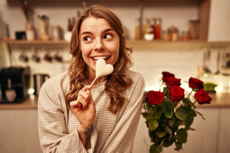 Foto de Feliz día de San Valentín. Una joven mujer feliz comiendo chocolate en forma de corazón mientras está sentada en una mesa en la cocina de su casa, con un ramo de rosas rojas cerca. - Imagen libre de derechos